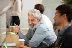 Older White Man Laughing With Younger Black Man At Desk With Takeaway Coffee